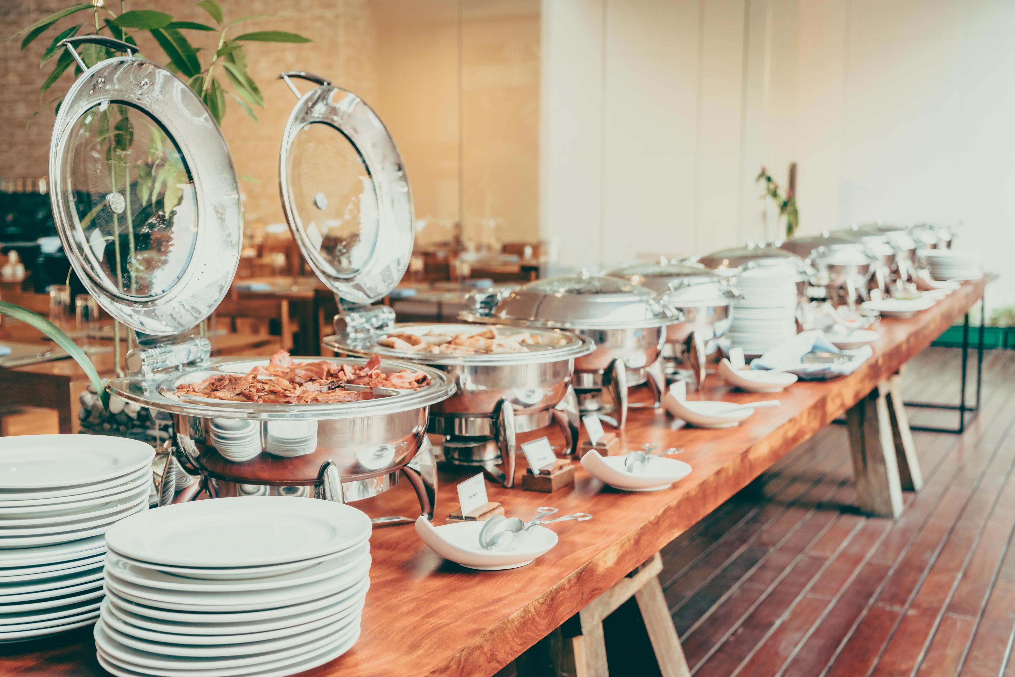 Buffet table with various chafing dishes, plates, and bowls set up in a bright room with wooden floors and plants.